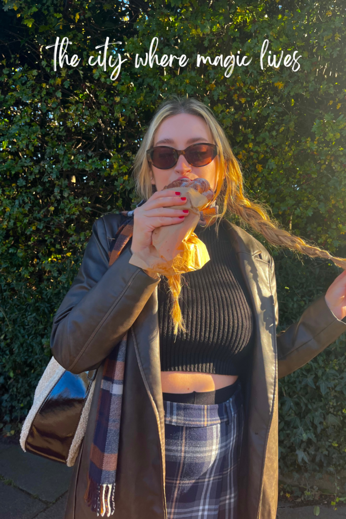 woman with plaits wearing sunglasses and a brown trench coat eating a pastry on dean bridge